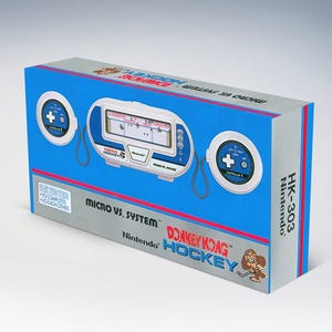 Könnte beinhalten: Eine blaue und silberne Nintendo Donkey Kong Hockey Micro Vs. System Spielkonsolenbox. Die Box zeigt eine weiße Spielkonsole mit einem Bildschirm und zwei kreisförmigen Controllern. Der Text "Donkey Kong Hockey" ist sichtbar.