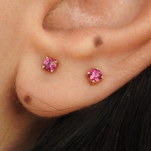 Peut inclure: Une paire de petites boucles d'oreilles en pierres précieuses roses avec des tiges en or. Les boucles d'oreilles sont portées dans les lobes d'oreille d'une personne aux cheveux foncés.