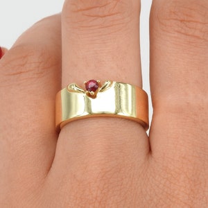 May include: A gold band ring with a red gemstone set in the center. The ring has a unique design with a curved band.