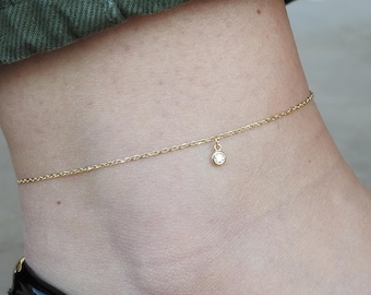 May include: A delicate gold anklet with a small, round, clear gemstone pendant. The anklet is worn on an ankle, with a glimpse of a black shoe and green fabric in the background.