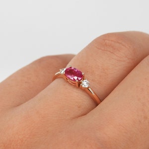 Puede incluir: Un delicado anillo de oro con una piedra ovalada rosa y dos piedras redondas blancas más pequeñas.