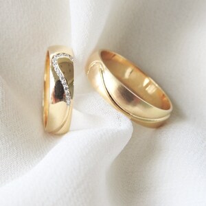 May include: Two gold wedding bands, one with a row of small diamonds, on a white fabric background.