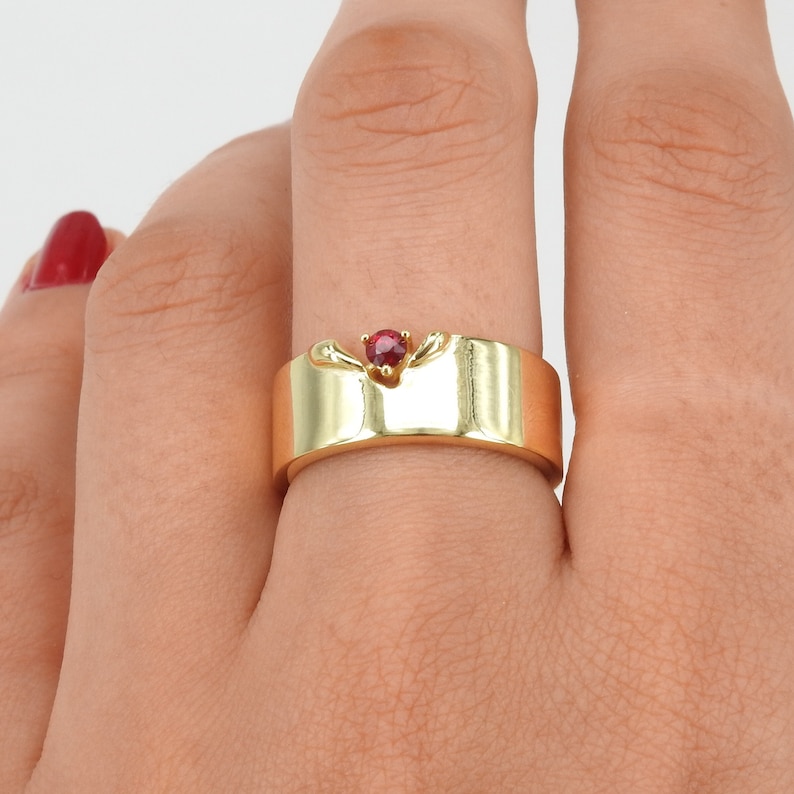 May include: A gold band ring with a red gemstone set in the center. The ring has a unique design with a curved band.