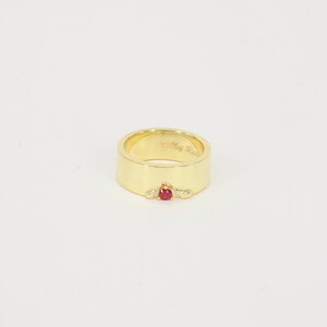 May include: A gold ring with a single red gemstone set in the center. The ring is wide and has a smooth, polished finish.
