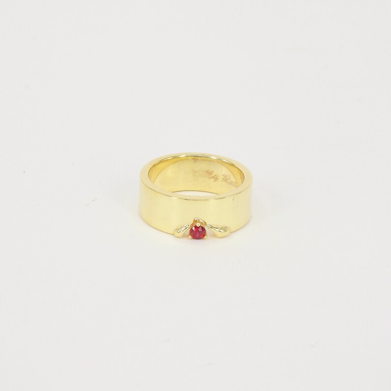 May include: A gold ring with a single red gemstone set in the center. The ring is wide and has a smooth, polished finish.