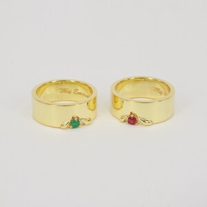 May include: Two gold wedding bands with a green emerald stone and a red ruby stone. The band on the left has the inscription "My Emerald".