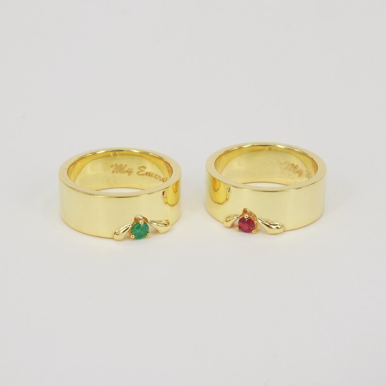 May include: Two gold wedding bands with a green emerald stone and a red ruby stone. The band on the left has the inscription "My Emerald".