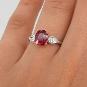 Puede incluir: Un anillo con una gran piedra de rubí de forma ovalada rodeada de dos diamantes en forma de pera. El anillo está engastado en oro blanco.