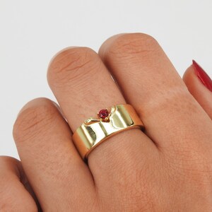 May include: A gold band ring with a red gemstone set in the center. The ring has a unique design with two small wings on either side of the stone.