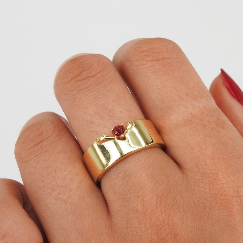 May include: A gold band ring with a red gemstone set in the center. The ring has a unique design with two small wings on either side of the stone.