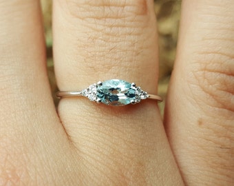 May include: A silver ring featuring a light blue oval gemstone, flanked by smaller clear stones on each side. The ring is set on a thin band, showcasing a delicate and elegant design. The ring is a piece of jewelry.