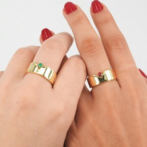 May include: Two gold rings with a green and red gemstone on each. The rings are on two hands, with the green ring on the left hand and the red ring on the right hand.