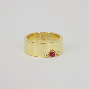 May include: A gold band ring with a single red gemstone set in a unique design.