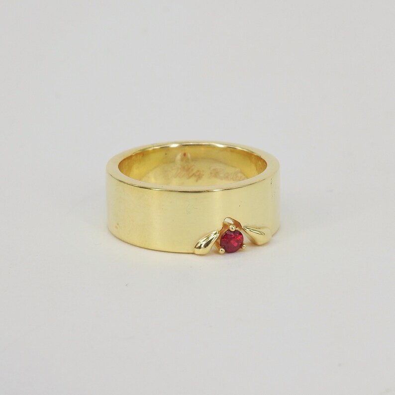 May include: A gold band ring with a single red gemstone set in a unique design.