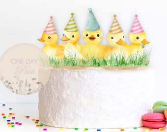 May include: A white cake with five yellow duck figurines wearing party hats. The ducks are arranged in a row on top of the cake. The cake is decorated with green grass and a brown base. The cake is on a white surface with colourful sprinkles scattered around it. A round, beige sticker with the text "ONE DAY Past" is on the left side of the cake.
