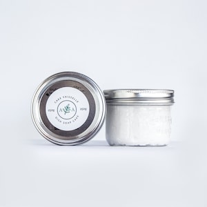 May include: Two silver jars with white lids. The jar on the left has a label that says "Cake Vaisselles 250g Dish Soap Cake 250g". The jar on the right is filled with a white substance.