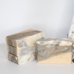 May include: A stack of three bars of handmade soap with a swirl pattern in shades of grey, white, and beige. A single bar of soap with a similar swirl pattern is in front of the stack. The soap has a logo with a leaf design and the word "Anya".