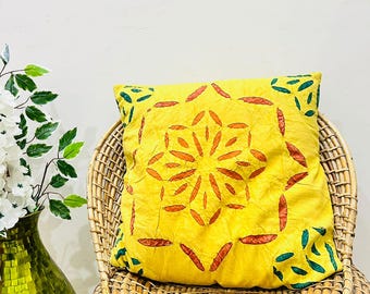 May include: A square yellow throw pillow featuring a central geometric pattern of orange leaf-shaped motifs arranged in a starburst design, with additional green leaf-shaped accents positioned near the four corners of the pillow cover.