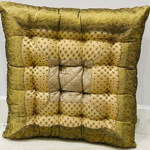Pode incluir: Almofada de chão quadrada e acolchoada em tons de dourado e verde-oliva. A almofada apresenta um quadrado central com acolchoado em losangos, rodeado por quadrados estampados com um desenho floral. As bordas são adornadas com um padrão tipo paisley. Aproximadamente 50 cm x 50 cm.