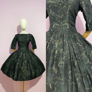 May include: A vintage black dress with a full skirt and elbow-length sleeves. The dress features a subtle gold pattern and is displayed on a mannequin. The dress has a boat neckline and a fitted bodice.