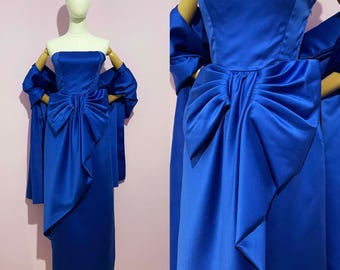 May include: A strapless, royal blue satin dress with a large bow detail at the waist. The dress is displayed on a mannequin and includes a matching shawl. The dress has a knee-length hemline and a fitted bodice.