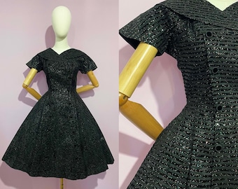May include: A vintage black dress with a textured, shimmering fabric. The dress features a collared neckline, short sleeves, and a flared skirt. The dress is displayed on a mannequin with wooden arms and legs.