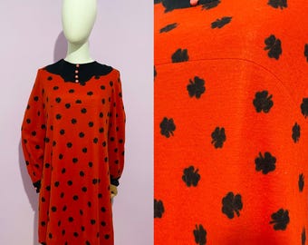 May include: A vintage-style, long-sleeved dress in a vibrant red hue, adorned with black clover-shaped patterns. The dress features a black collar with pink buttons and black cuffs. The dress is displayed on a mannequin.