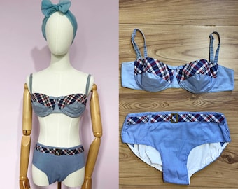 May include: A light blue bikini set with a plaid pattern in red, white, and navy blue. The set includes a bra, high-waisted bottoms, and a matching headband. The bra and bottoms have a belt detail.