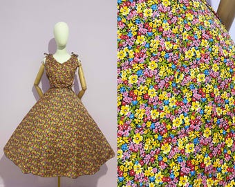 May include: A vintage-style dress with a floral pattern, displayed on a mannequin. The dress features a V-neckline, tie straps, and a fitted bodice that flares into a full skirt. The fabric is a mix of yellow, pink, red, and blue flowers on a brown background.