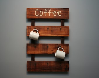 May include: Wooden coffee mug rack with the word "Coffee" in cursive script. The rack has four horizontal shelves with hooks for mugs, and two white mugs are hanging on the hooks. The rack is made of dark brown wood.