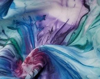 May include: Close-up of a flowing fabric with a watercolor effect. The fabric features a blend of blues, purples, and greens, with a marbled or swirling pattern. The colors blend and create a soft, artistic look. The fabric appears to be lightweight and possibly silk.