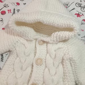 Puede incluir: Primer plano de un cárdigan blanco de punto para bebé con capucha. El cárdigan presenta un diseño de punto trenzado en la parte delantera y botones de madera. La capucha también está tejida. El fondo es una tela estampada con ilustraciones.