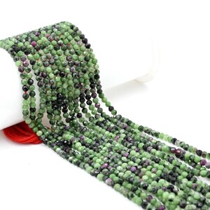 Può includere: Fili di perline di pietre preziose sfaccettate nei toni del verde, nero e rosso. Le perline sono piccole e infilate insieme, creando un aspetto fluido e strutturato. Le pietre preziose sono esposte su uno sfondo bianco.