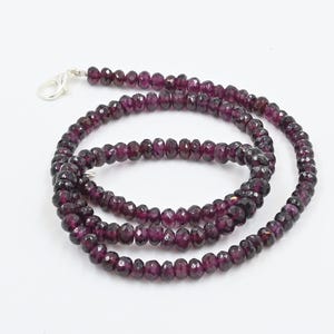 May include: A necklace composed of small, faceted, deep purple beads. The beads are strung together, forming a continuous strand that curves and coils. A silver-colored clasp secures the necklace.