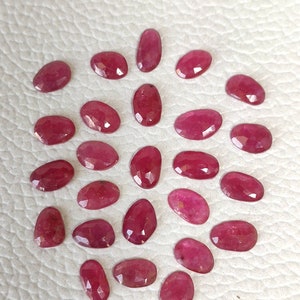 May include: A collection of 25 loose, faceted, oval-shaped ruby gemstones. The rubies are a deep red color and have a polished, shiny finish.