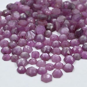 Puede incluir: Una colección de gemas facetadas de color rosa púrpura. Las gemas varían en tamaño y forma, con una mezcla de formas hexagonales e irregulares. Las gemas están dispersas sobre una superficie blanca, creando una exhibición visualmente atractiva de color y luz.