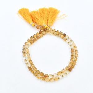 Puede incluir: Una hebra de cuentas cuadradas de color marrón claro y transparentes con detalles dorados. Las cuentas están enhebradas en un cordón transparente y tienen un borla en cada extremo.