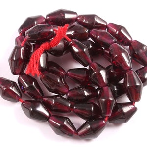 Puede incluir: Una hebra de cuentas de piedras preciosas facetadas de color rojo oscuro. Las cuentas son todas de tamaño y forma similares, y están enhebradas en un hilo rojo fino. Las cuentas están dispuestas en un patrón suelto y fluido.