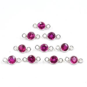 Op de afbeelding: Een verzameling van tien zilverkleurige connectoren, elk met een levendige magenta edelsteen. De ronde edelstenen zijn gezet in een bezel setting. De connectoren hebben kleine lussen aan elk uiteinde, perfect voor het maken van sieraden.