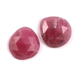 May include: Two faceted, heart-shaped gemstones in a deep pink hue. The gems have multiple facets that catch the light, creating a sparkling effect. The stones are set against a white background.