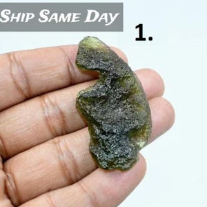 May include: A rough, olive-green gemstone with a textured surface is held in a hand. The stone has a natural, irregular shape. The text "SHIP SAME DAY" is visible in the upper left corner, and the number "1." is in the upper right corner.