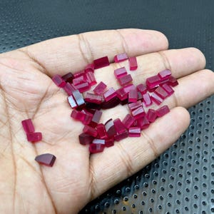 May include: A collection of small, rectangular ruby gemstones in varying shades of red and pink. The gemstones are held in a hand, showcasing their faceted surfaces and natural color variations. The gemstones are raw and uncut.