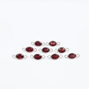 May include: A collection of oval-shaped, deep red gemstones set in silver-colored metal connectors. Each gem is displayed against a white background. The connectors have small loops on each end, suggesting they are designed for jewelry making.