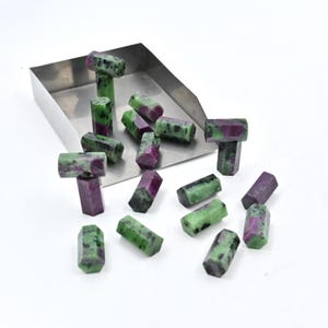 Può includere: Una collezione di pietre grezze di gemme verdi e viola di diverse forme e dimensioni. Le pietre grezze sono disposte su un vassoio di metallo e una superficie bianca.