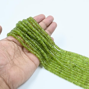 May include: Strands of light green peridot gemstone beads. The beads are small, square-cut, and strung together. The beads are held in a hand against a white background. The color is a vibrant, translucent green.