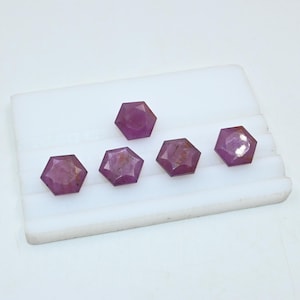May include: Five pink, faceted, hexagonal gemstones on a white surface.