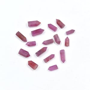 Puede incluir: Una colección de puntas de cristal de rubí rosa, cada una con una punta puntiaguda y una superficie lisa y pulida. Los cristales están dispersos sobre un fondo blanco.