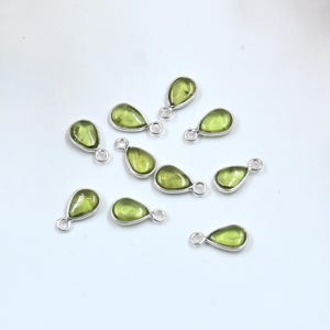 Könnte beinhalten: Eine Sammlung tropfenförmiger Anhänger mit einem hellgrünen Edelstein in der Mitte und einem silberfarbenen Metallrand. Jeder Anhänger hat oben eine kleine Schlaufe zum Befestigen an Schmuck. Die Anhänger sind auf einer weißen Oberfläche verstreut.