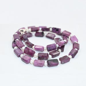 Op de afbeelding: Een ketting met rechthoekige, dieppaarse edelstenen afgewisseld met kleine, witte parels. De edelstenen hebben een facetgeslepen uiterlijk en de ketting is vastgezet met een zilveren sluiting. De sieraden worden gepresenteerd tegen een witte achtergrond.