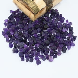 May include: A large pile of small, rough, purple amethyst crystals. The crystals are scattered on a white surface.